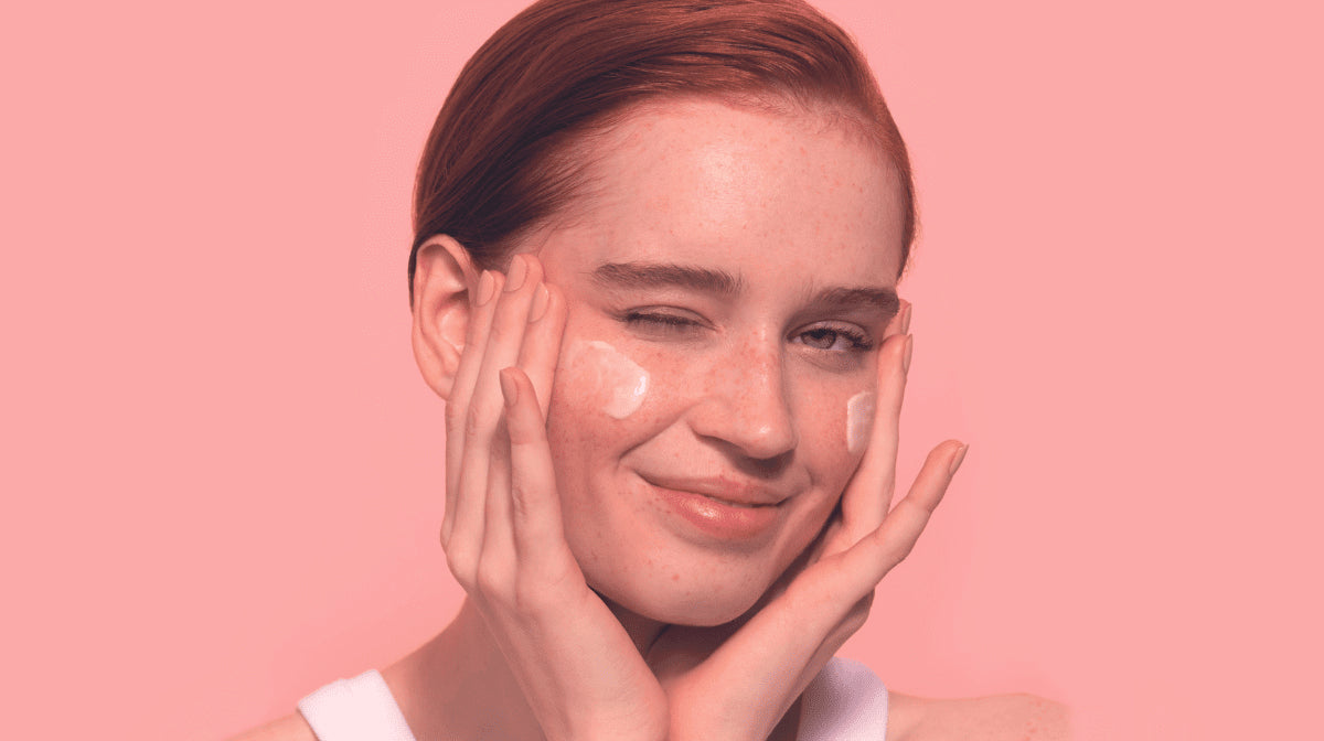 A woman with clear, glowing skin and skincare cream on her face winks and smiles at the camera